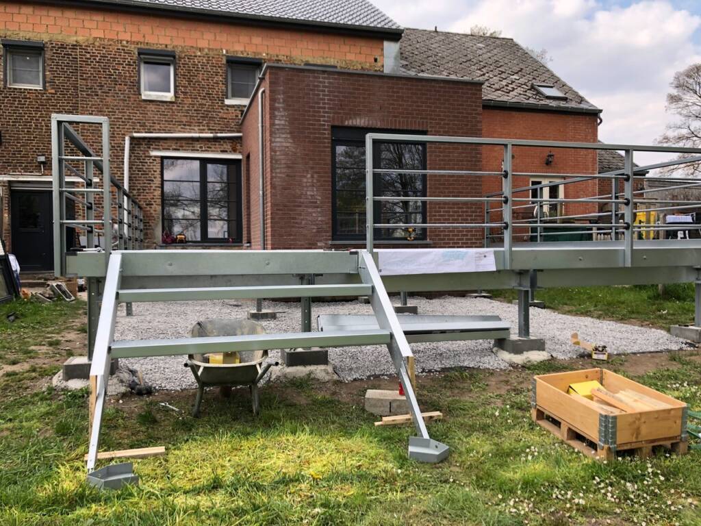 Montage d’une ossature métallique ModuHome pour terrasse sur pilotis attenante à une maison en briques.